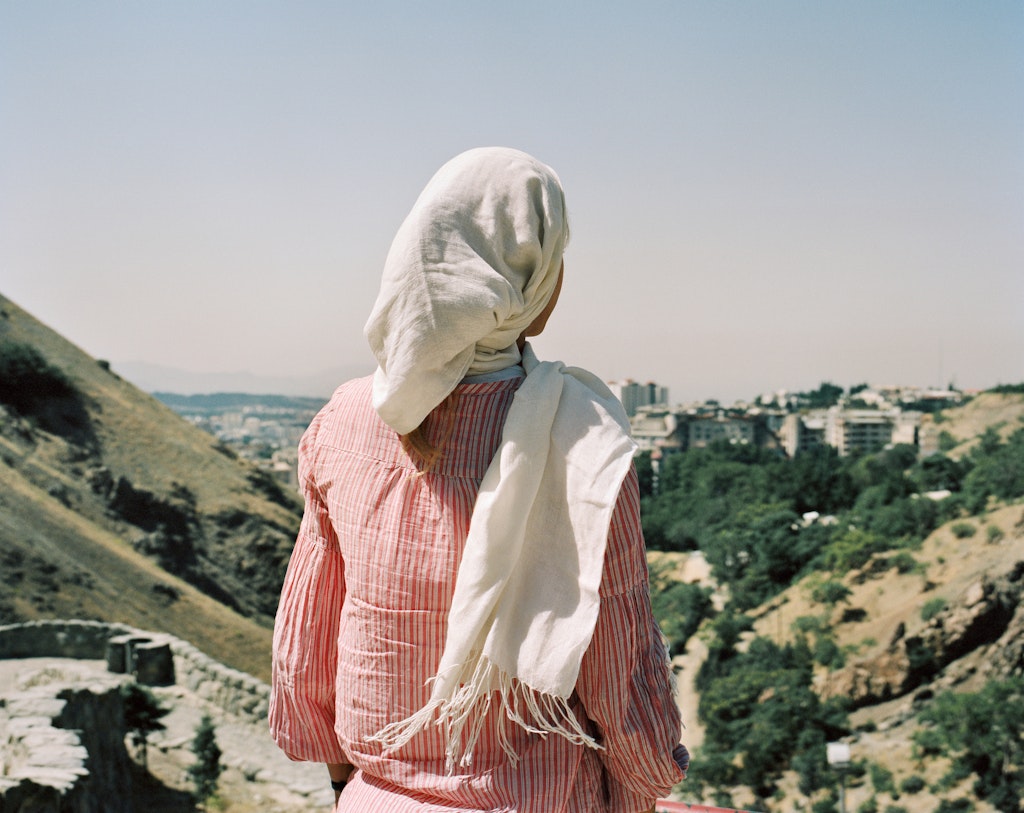 The back of a person with a long scarf around their head, looking over grassy hills to distant trees and city buildings