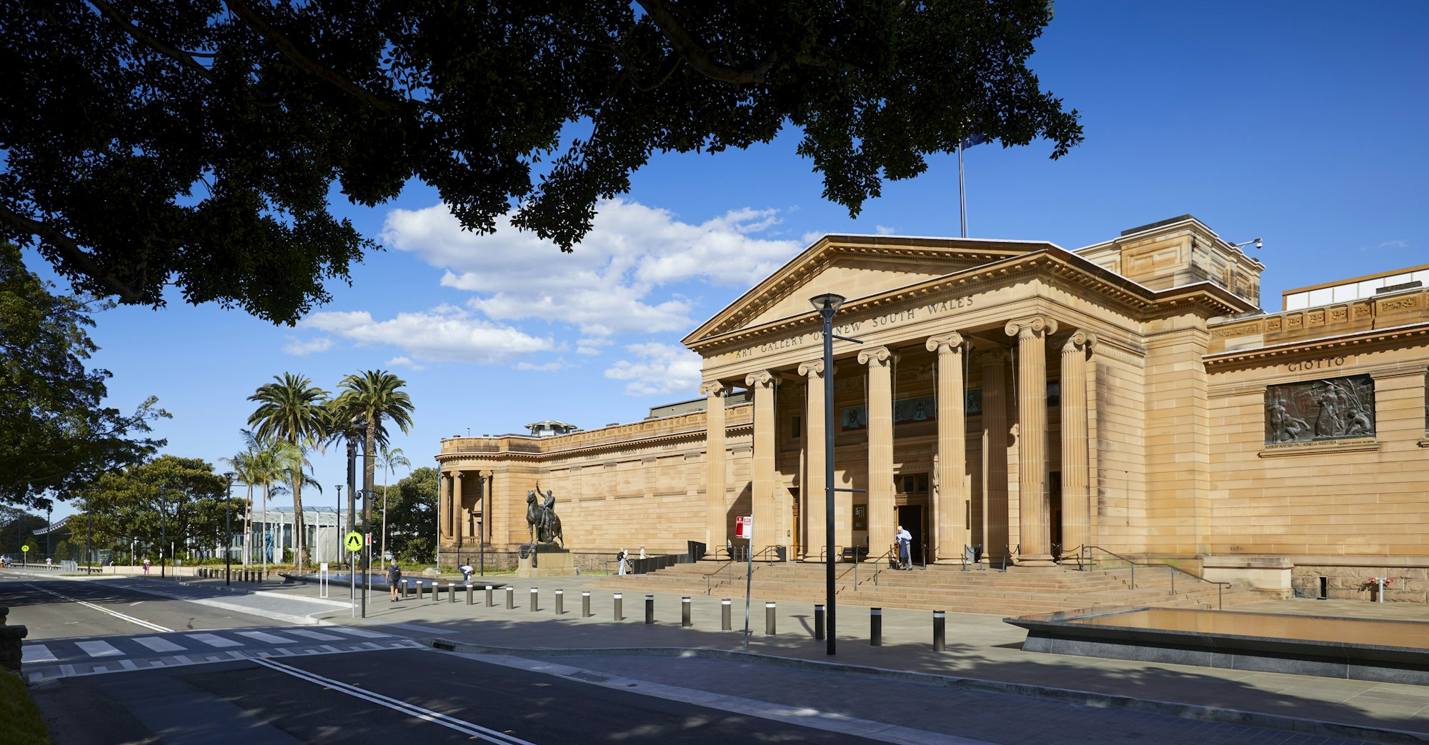 View across a street of a large sandstone building with a portico and more distant trees and building