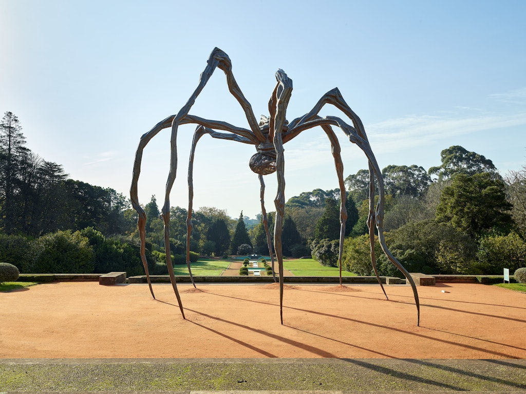 A ten-metre tall bronze sculpture of a spider installed on a sandy surface. In the distance is a garden lined with trees.