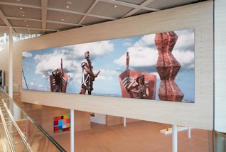 A large video screen showing a standing figure against a sky with two figures seated in boats