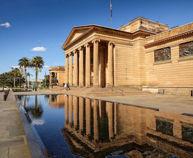 A sandstone building with a columned portico is reflected in a decorative pool