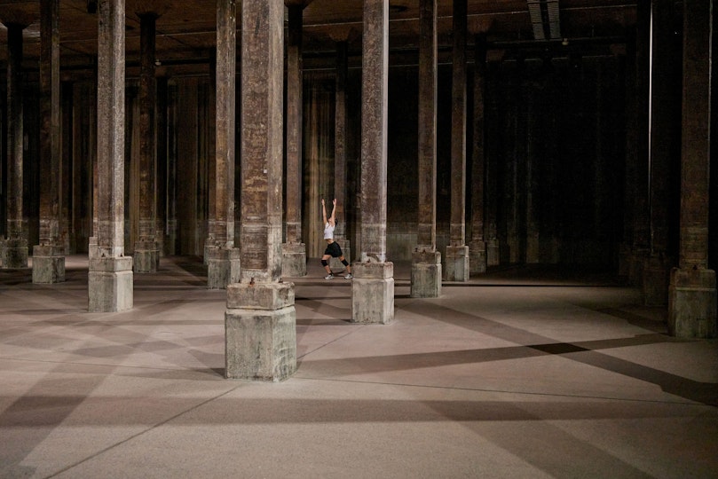 A person lunges with their arms stretched upwards in a shadowy columned space