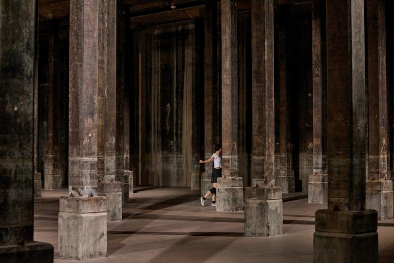 A person stands with an arm held out sideways and a leg bent in a shadowy columned space