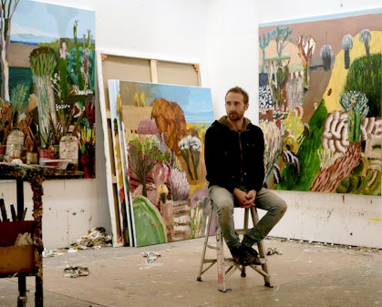 A person sits on a stool with large paintings around them and discarded paint tubes on the floor
