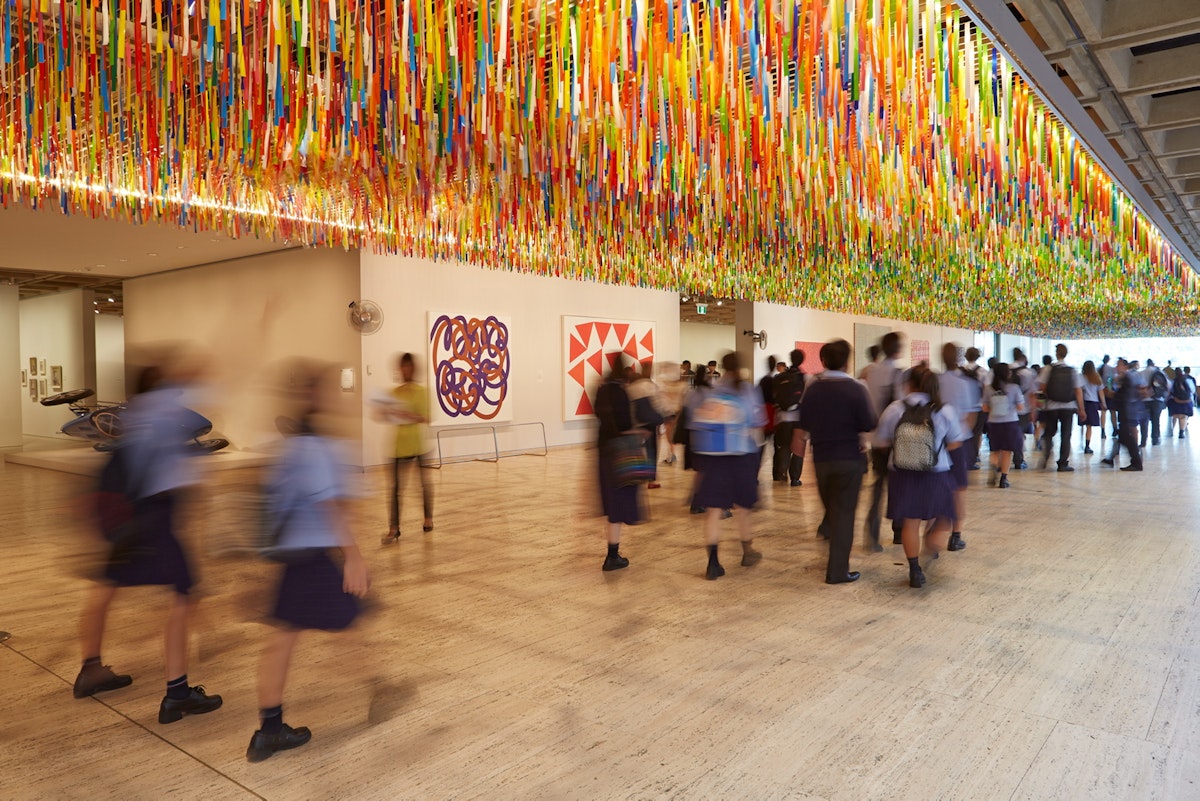 A large group of high school students walking under an artwork made of colourful plastic streamers suspended from the ceiling.