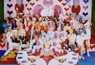 A large group of people in colourful costumes pose for a group photograph in a setting decorated with hearts and dots