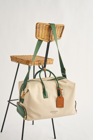 photo of a large cream weekender bag with green trim handing on the back of a wicker chair