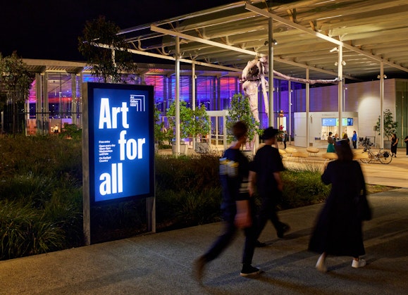 Three people walk past an illuminated sign saying 'Art for all' towards a glass-fronted building under a large awning