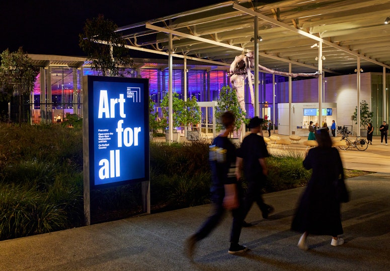 Three people walk past an illuminated sign saying 'Art for all' towards a glass-fronted building under a large awning