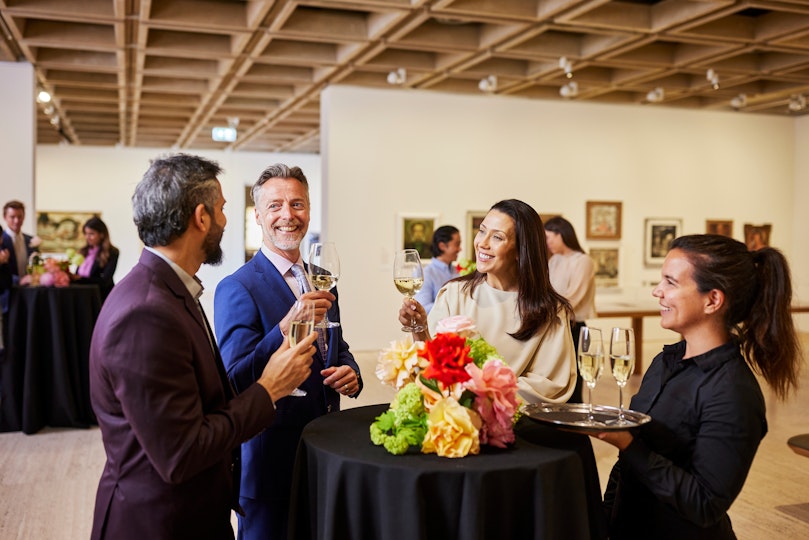 Four people holding wine standing around a table with flowers in a Gallery space with paintings on the walls behind them.