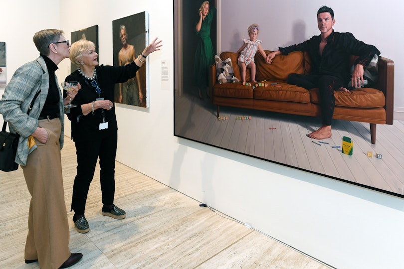 Two people looking at a painting that shows a women in a green dress standing to the far left and in shadow, a toddler with blonde curls standing on a couch and a man in black jeans and shirt sitting on the couch. Blocks and crayons are scattered on the couch and floor.