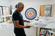 A person stands holding an open notebook. Behind them is a large artwork of concentric circles and around them are tables holding art materials.