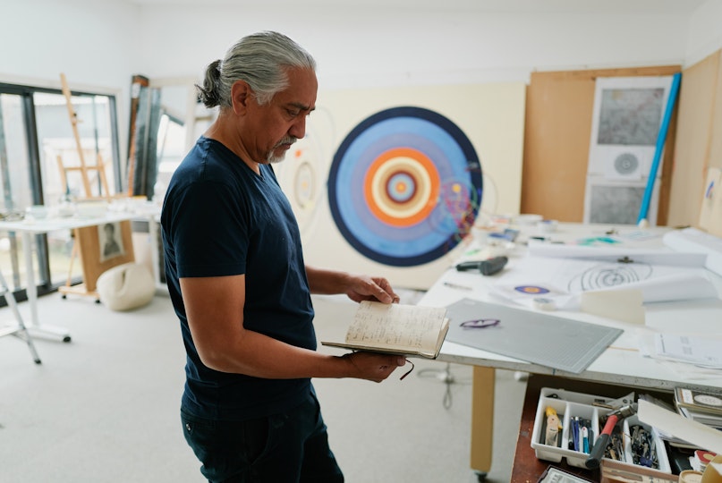 A person stands holding an open notebook. Behind them is a large artwork of concentric circles and around them are tables holding art materials.