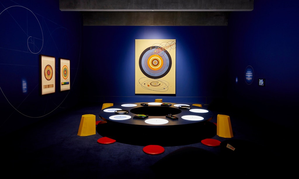 A dark blue room with geometric artworks hung on the walls and a central circular table and bench