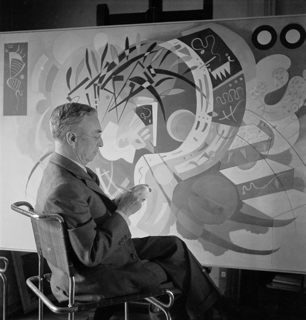 A man with short hair and glasses wearing a suit sits with his legs crossed in front of a large painting.