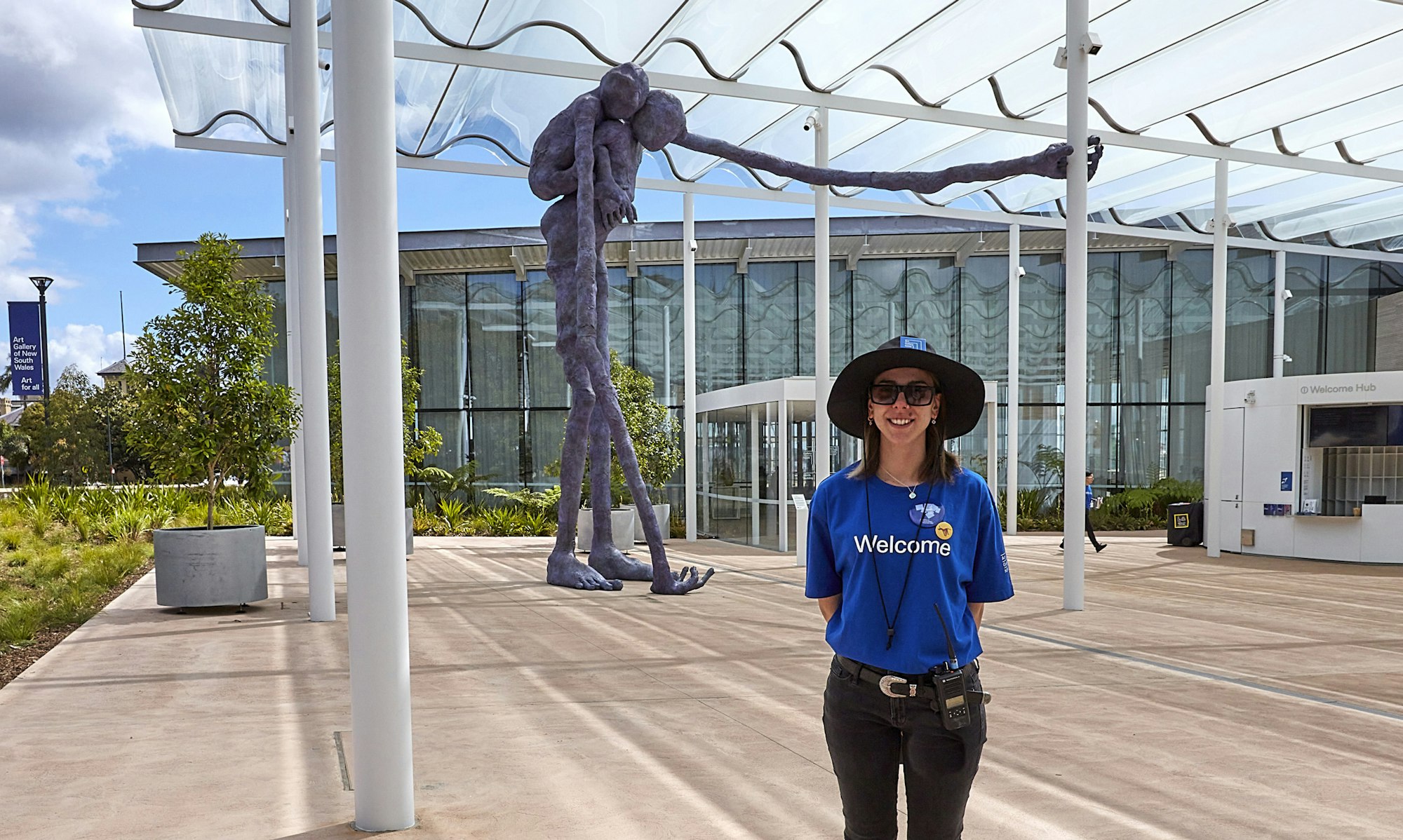 A person wearing a t-shirt with the word 'Welcome' stands in front of a huge purple figure sculpture outside a building
