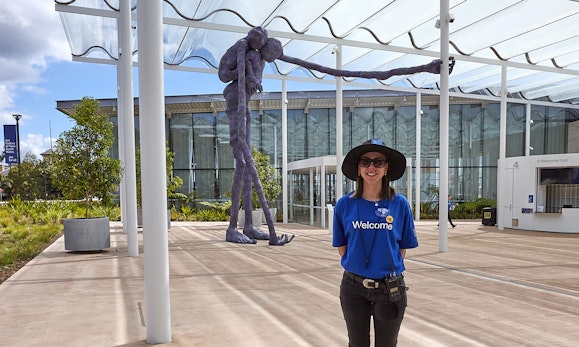 A person wearing a t-shirt with the word 'Welcome' stands in front of a huge purple figure sculpture outside a building