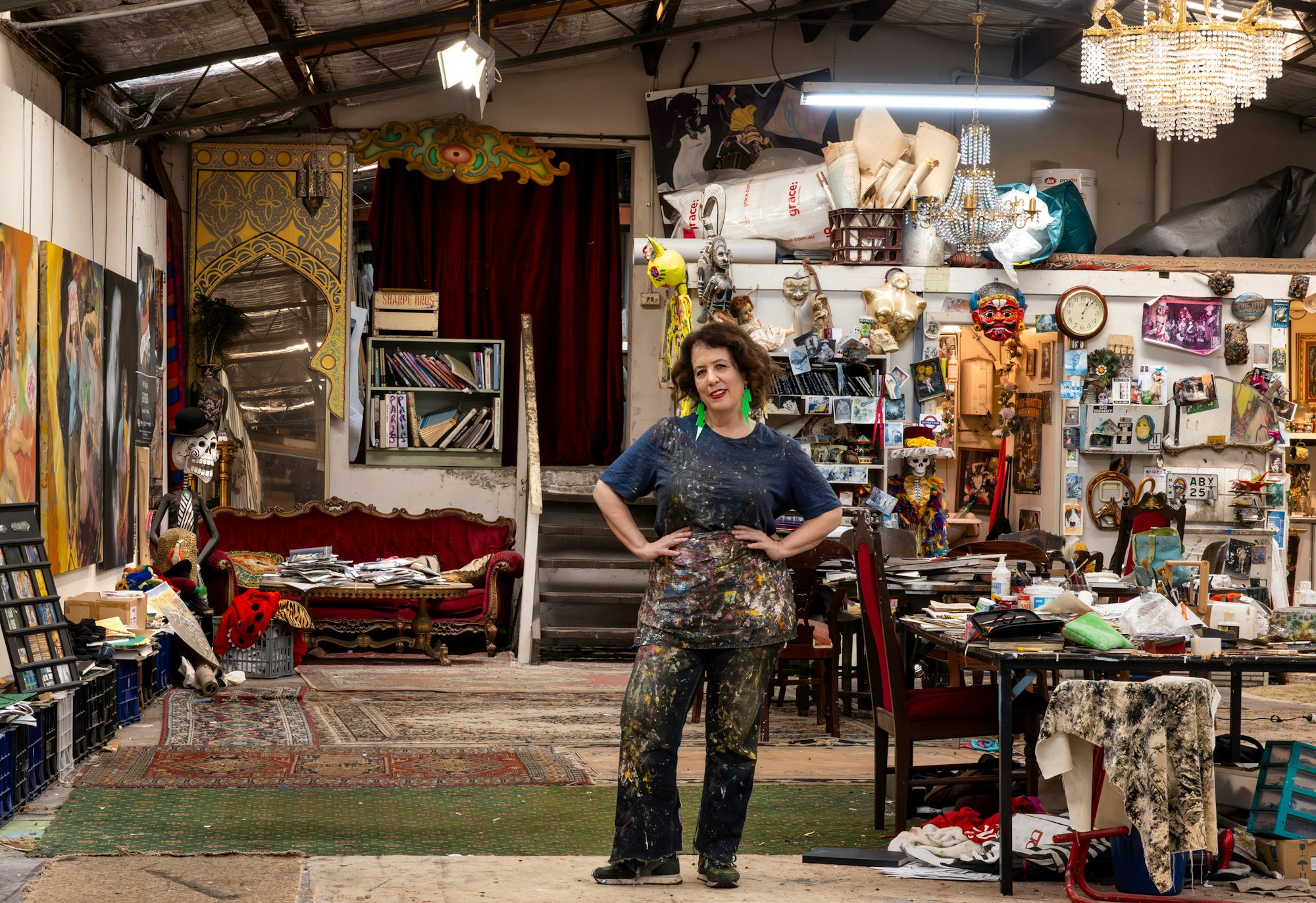 A person in paint-spattered clothes stands in a large high-ceilinged room surrounded by many art-related objects