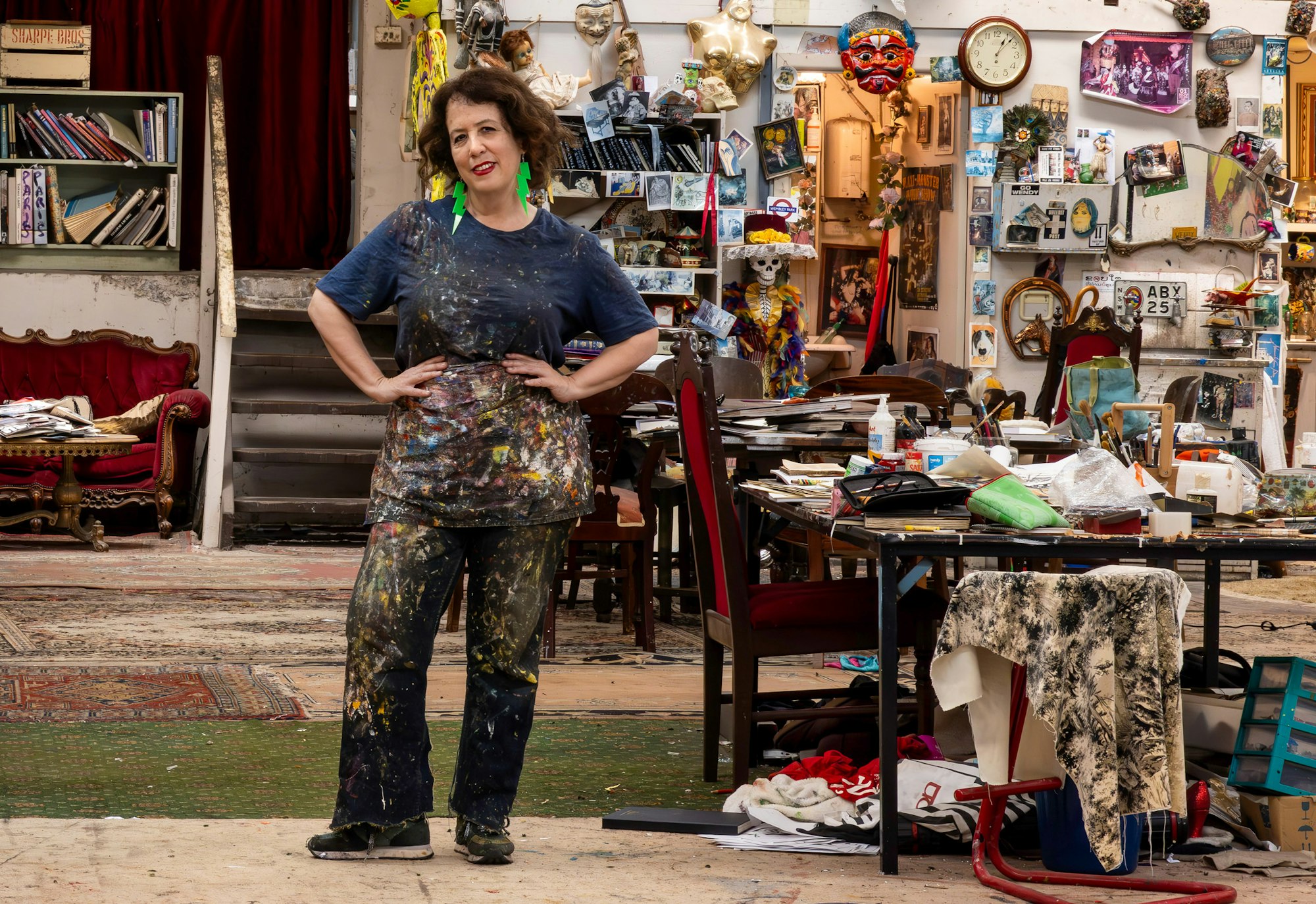 A person in paint-spattered clothes stands surrounded by many art-related objects