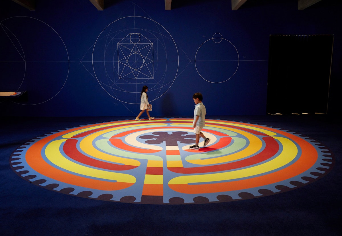 Two children walk on a coloured labyrinth in a dark room with geometric patterns on the wall
