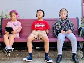 Three children, wearing headphones, sit on a bench