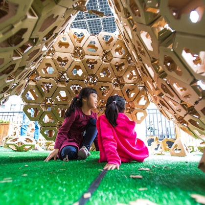 Two children sit on the ground under a cardboard structure