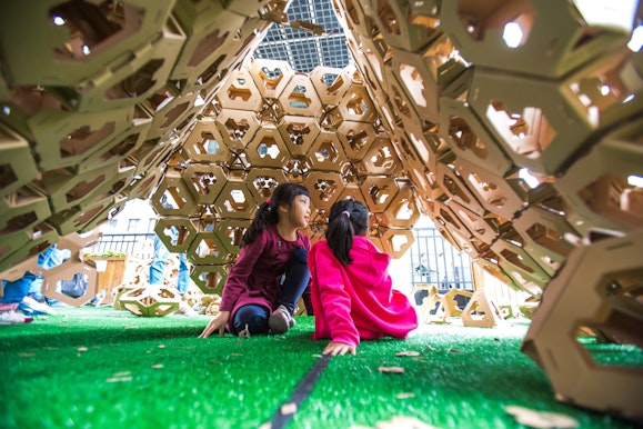 Two children sit on the ground under a cardboard structure