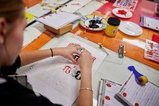 A person works on a text-based artwork at a table strewn with notepads and art materials