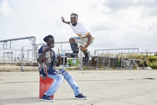 One sitting person and one jumping person outdoors beside industrial fencing and train wires