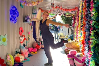 A person stands on one leg in a shed full of colourful plush objects and textile hangings
