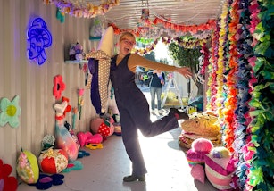 A person stands on one leg in a shed full of colourful plush objects and textile hangings