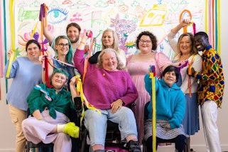 A group of 10 people holding long coloured ribbons in front of a colourful wall drawing