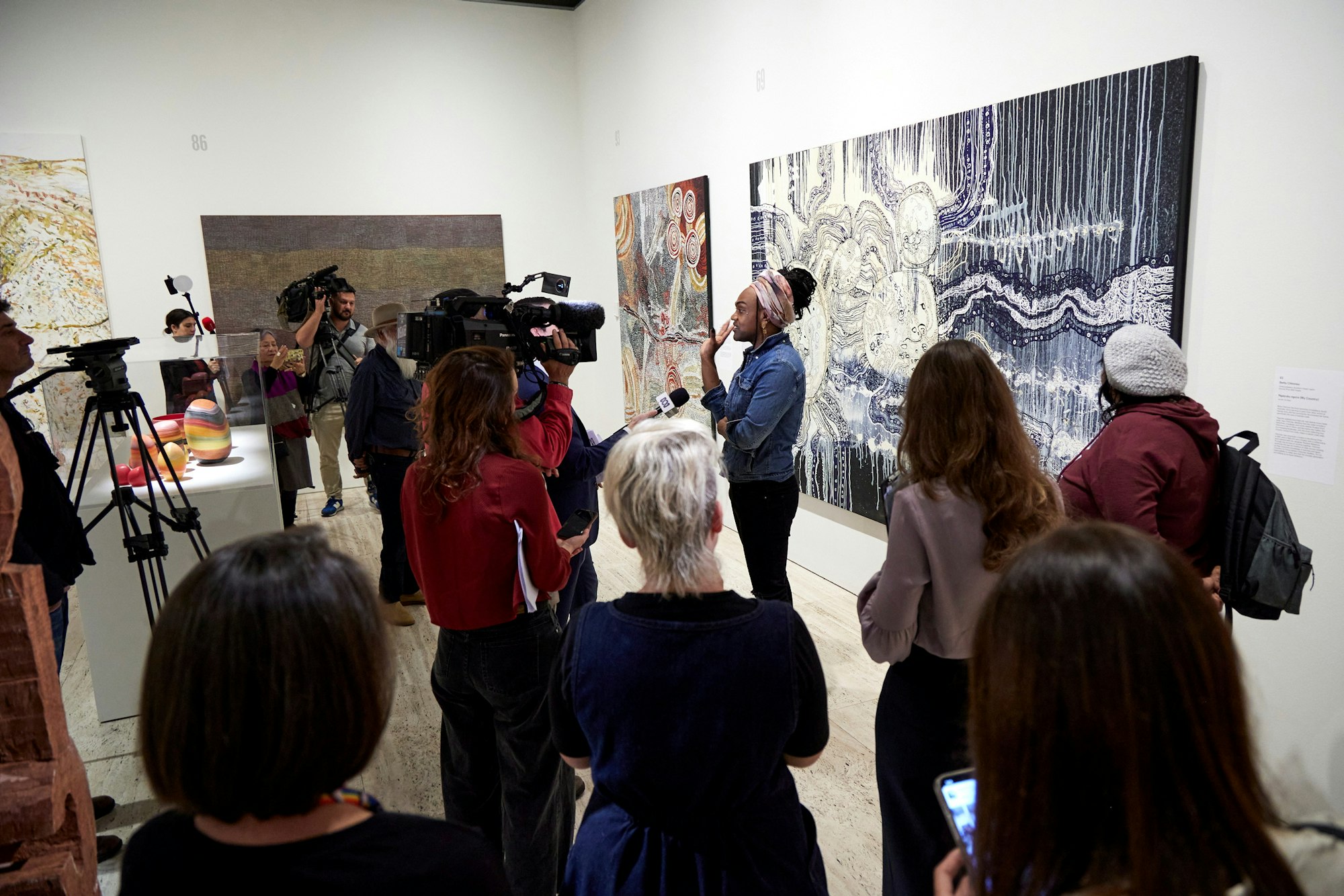 A person stands with a large black and white painting in front of a group of people, some with cameras