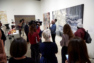 A person stands with a large black and white painting in front of a group of people, some with cameras