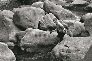 A black-and-white photo of a person standing among large boulders, holding a paintbrush above a series of painted marks on one boulder