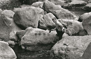 A black-and-white photo of a person standing among large boulders, holding a paintbrush above a series of painted marks on one boulder