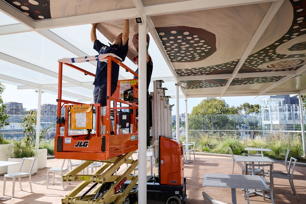 Two people on a scissor lift underneath an artwork on an outdoor canopy above tables