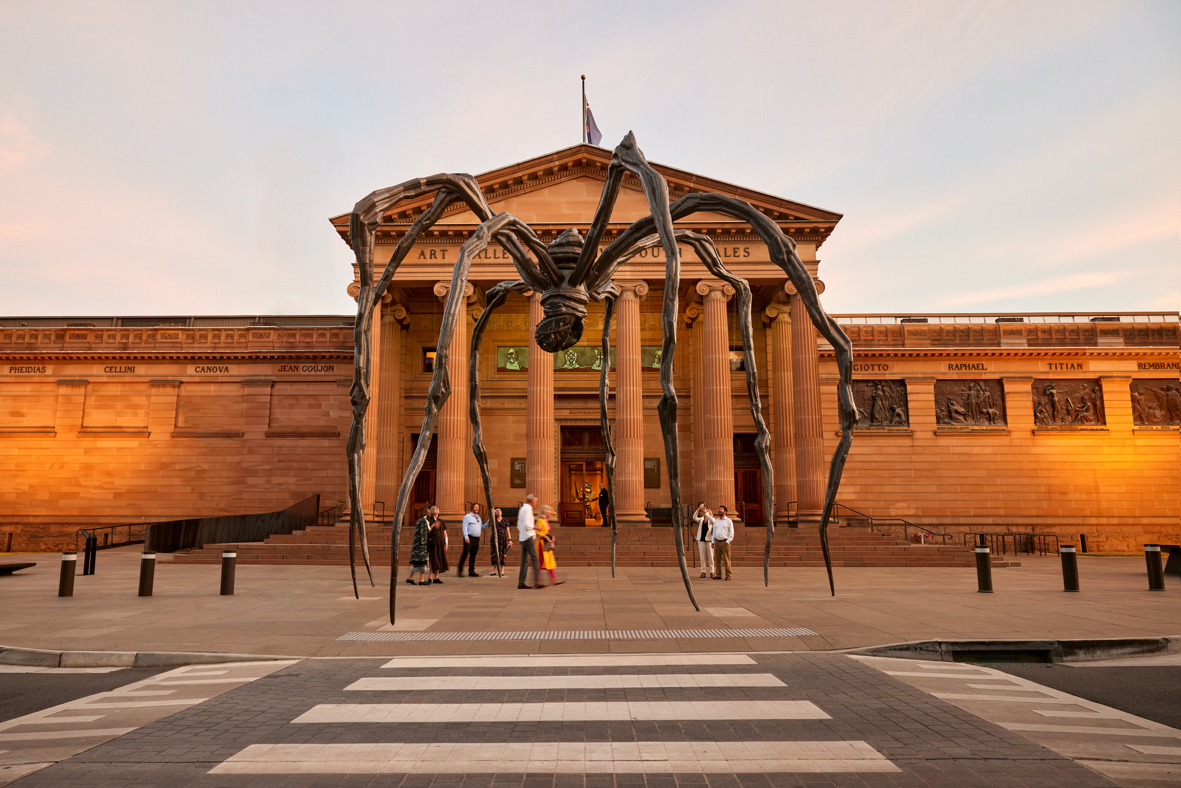 A giant spider sculpture in front of a grand sandstone building with Art Gallery of New South Wales above the portico at sunset.