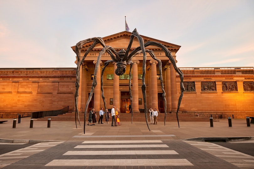 A giant spider sculpture in front of a grand sandstone building with Art Gallery of New South Wales above the portico at sunset.