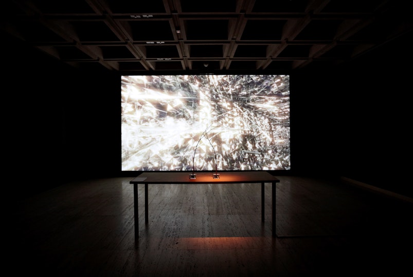 A dark gallery space with a bright abstract image on a large screen that has a table and two joysticks in front of it