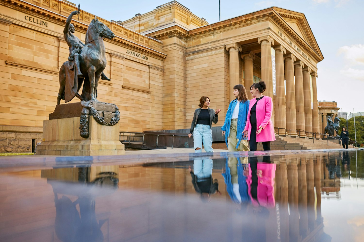 Three people stand at the front of a large sandstone building next to a sculpture of a person on a horse