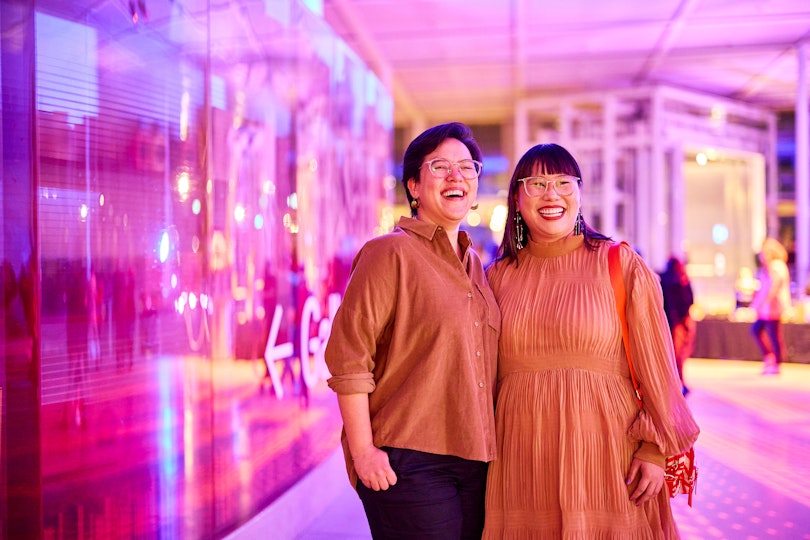 Two people with black hair and glasses and dressed in orange in front of a pink-lit building