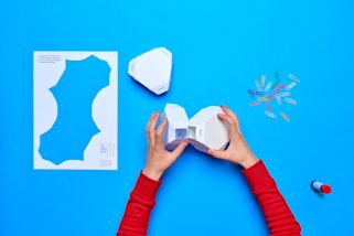 Two hands holding two geometric 3D cardboard shapes. Nearby is another shape, a piece of paper that has been cut out, a pile of paperclips and a glue stick