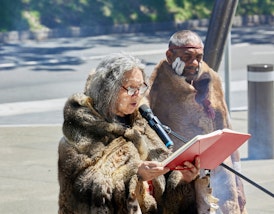 Two people wearing fur cloaks. The person on the left holds a red book and speaks into a microphone.