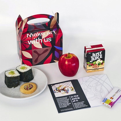 A handled box with words 'Make art with us', a Just Juice container, apple, sushi rolls and a cookie on a plate, booklet and small box of pencils