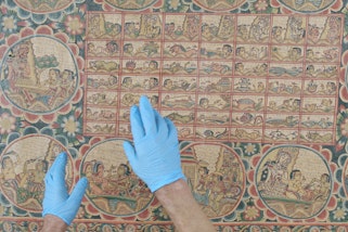 Two gloved hands above a textile artwork depicting various scenes