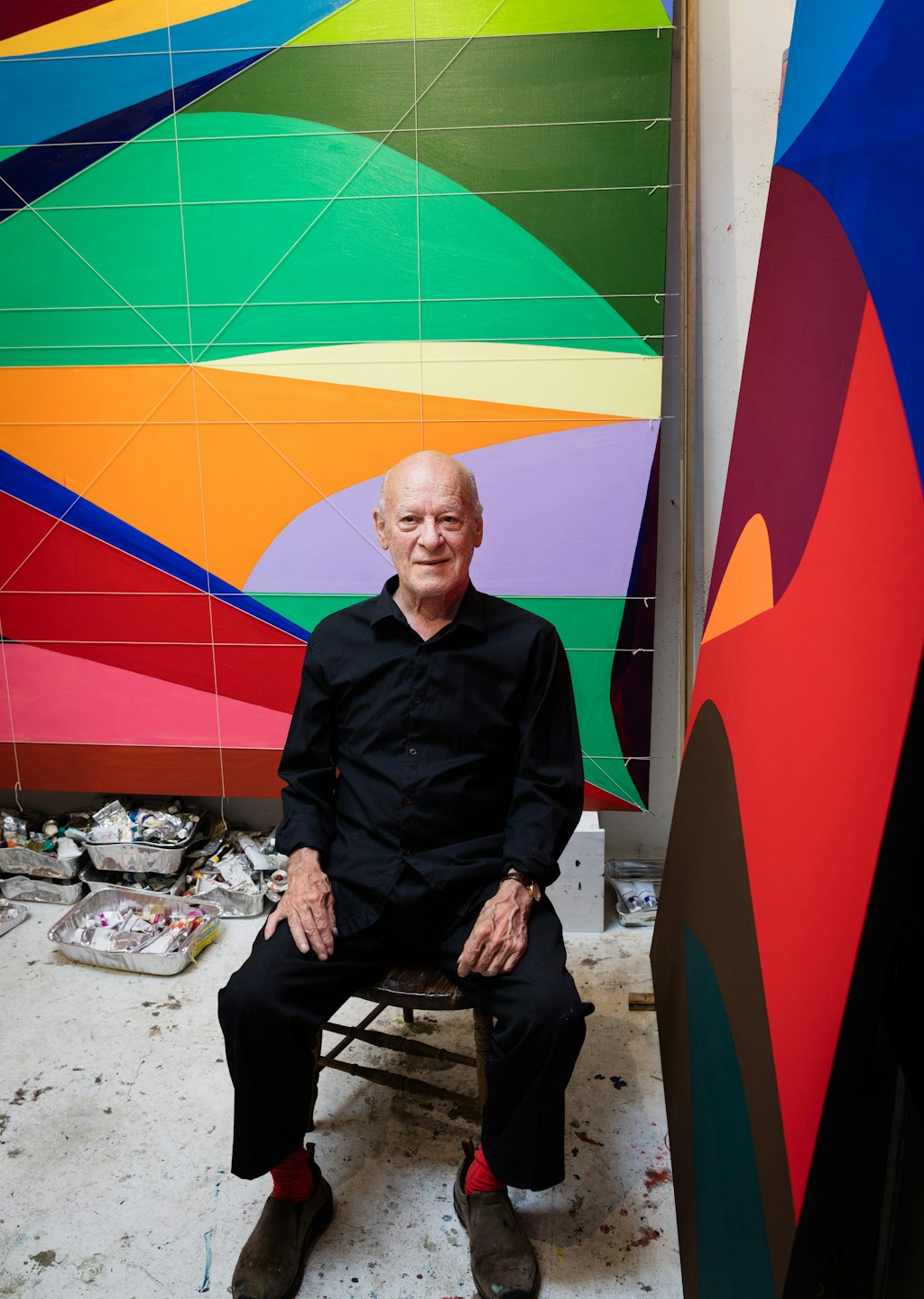 A person dressed in black sits between two large paintings of colourful abstract shapes. Art materials are on the floor.