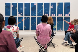 A small group of people sit on portable stools in front of an artwork in a gallery space