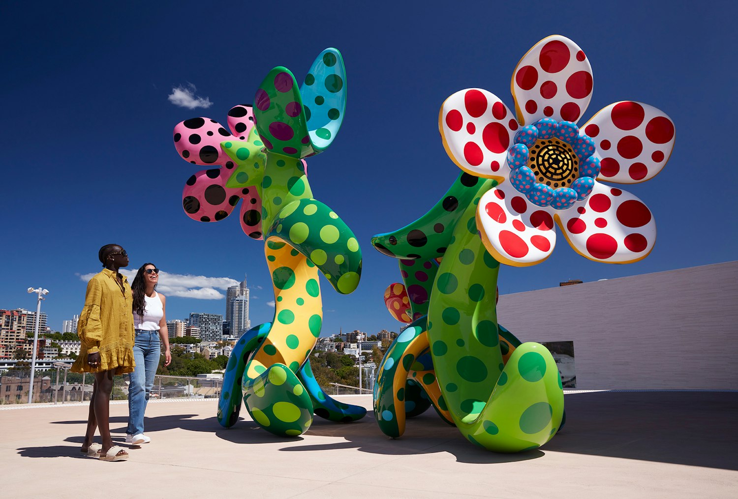 Two people on a terrace outdoors next to a large spotted flower sculpture with a city skyline in the background