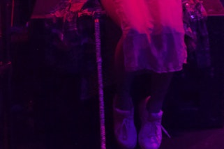 A pair of legs, dangling off the edge of a black box, covered to just below the knees by a sheer slip dress. white trainers on feet. Beside the legs, resting against the box that the person sits on is a stripey cane, with dark handle. The image is awash in pink and purple light.